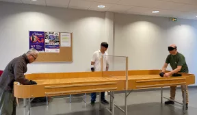 Photo d'une table de showdown , la table est longue en bois clair et dispose de rebord  tout le long de la tableau. Au centre est installé sur toute da largeur un panneau en plexi glace maintenu à 10 centimètre au dessus de la table. Au deux extrémités deux joueurs et au milieu un arbitre. 