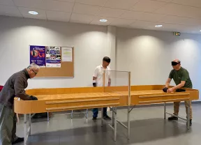 Photo d'une table de showdown , la table est longue en bois clair et dispose de rebord  tout le long de la tableau. Au centre est installé sur toute da largeur un panneau en plexi glace maintenu à 10 centimètre au dessus de la table. Au deux extrémités deux joueurs et au milieu un arbitre. 