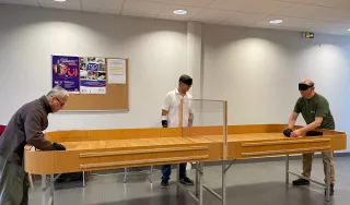 Photo d'une table de showdown , la table est longue en bois clair et dispose de rebord tout le long de la tableau. Au centre est installé sur toute da largeur un panneau en plexi glace maintenu à 10 centimètre au dessus de la table. Au deux extrémités deux joueurs et au milieu un arbitre.