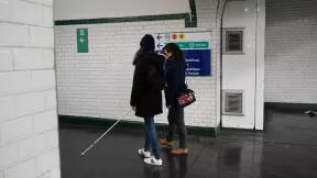 Une personne déficiente visuelle et son accompagnatrice en train de se déplacer dans les couloirs du métro parisien.