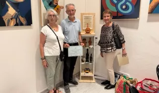 L’image montre deux femmes et un homme debout dans un cadre intérieur, dans un espace d’exposition. - Sur le mur derrière le groupe, il y a deux grandes œuvres d’art. Celle de droite présente un motif avec des formes circulaires imbriquées, tandis que celle de gauche semble représenter des figures abstraites. - Au centre de l’arrière-plan, il y a une étagère d’exposition en bois contenant plusieurs petites sculptures et artefacts, dont un buste et d’autres figurines.