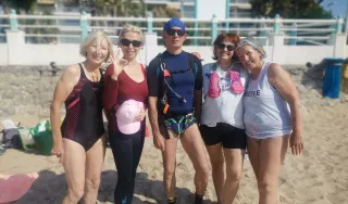 Cinq personnes debout près les uns des autres sur une plage de sable. Ils sont positionnés sur une seule rangée, les bras les uns autour des autres, suggérant une pose de groupe.