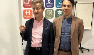 Sylvain Nivard, président de l'association Valentin Haüy (à gauche), et le directeur de la fédération (à droite) posent côte à côte devant un kakémono d'information. Sylvain Nivard, souriant, porte une veste bleu marine sur une chemise à rayures roses et tient une canne blanche. Le directeur porte un costume marron clair sur une chemise bleue. Le panneau derrière eux présente le logo de l'association « Valentin Haüy : Agir pour l'autonomie » ainsi que des icônes colorées illustrant leurs services : médiathè
