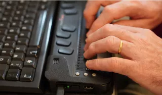 Gros plan sur les mains d’une personne utilisant une plage braille connecté à un clavier d’ordinateur.