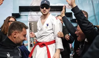 Nohan Dudon, athlète de karaté, portant un kimono blanc avec un écusson de l'équipe de France et une ceinture rouge, se tient debout au centre de l'image. Il porte également une casquette bleu marine et des lunettes de protection teintées. Il est entouré de plusieurs personnes qui l'acclament, les mains levées en signe de célébration. À l'arrière-plan, on aperçoit une affiche pour le « 27th Karate World Championships » au Caire.