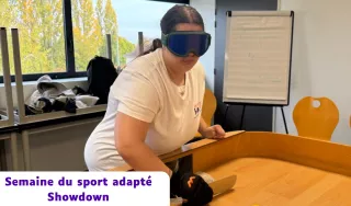 Une femme portant un masque occultant bleu et un gant de protection participe à une partie de Showdown lors de la Semaine du sport adapté. Elle est penchée au-dessus d'une table de jeu en bois rectangulaire avec des bords surélevés, tenant une raquette plate pour diriger une balle sonore. En arrière-plan, on aperçoit une salle lumineuse avec un tableau de conférence et des chaises en bois.