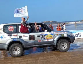 Durant la parade d'arrivée à Essaouira, Laure assise sur la portière arrière droite , agite le drapeau de l'association. En arrière-plan : la plage d'Essaouira.