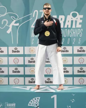 Nohan Dudon debout sur la première marche du podium, célébrant sa médaille d'or aux Championnats du Monde de Karaté 2025 au Caire. Il porte un pantalon de kimono blanc, une veste de survêtement bleu marine de l'équipe de France et des lunettes de protection teintées. Il a la main droite posée sur le cœur devant un mur de logos incluant "Daedo" et l'emblème de la compétition.