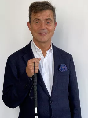 Portrait à mi-corps de Sylvain Nivard, président de l'association Valentin Haüy. Il sourit face à l'objectif, vêtu d'un blazer bleu marine avec une pochette à motifs et d'une chemise blanche ouverte au col. D'une main, il tient la poignée de sa canne blanche repliée. Le fond est neutre et de couleur claire.