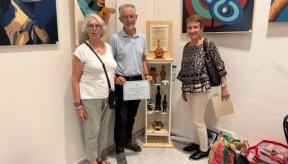 L’image montre deux femmes et un homme debout dans un cadre intérieur, dans un espace d’exposition. - Sur le mur derrière le groupe, il y a deux grandes œuvres d’art. Celle de droite présente un motif avec des formes circulaires imbriquées, tandis que celle de gauche semble représenter des figures abstraites. - Au centre de l’arrière-plan, il y a une étagère d’exposition en bois contenant plusieurs petites sculptures et artefacts, dont un buste et d’autres figurines.