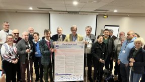 Une vingtaine de personnes sont réunies en groupe dans une salle de conférence pour la signature de la « Charte Romain Jacob ». Au centre, plusieurs hommes tiennent une grande affiche blanche détaillant les points de la charte, qui vise à améliorer l'accès aux soins des personnes en situation de handicap. Plusieurs membres du groupe tiennent des cannes blanches et une femme au premier plan est accompagnée d'un chien guide noir. L'ambiance est formelle et chaleureuse, marquée par de nombreux sourires.