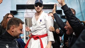 Nohan Dudon, athlète de karaté, portant un kimono blanc avec un écusson de l'équipe de France et une ceinture rouge, se tient debout au centre de l'image. Il porte également une casquette bleu marine et des lunettes de protection teintées. Il est entouré de plusieurs personnes qui l'acclament, les mains levées en signe de célébration. À l'arrière-plan, on aperçoit une affiche pour le « 27th Karate World Championships » au Caire.