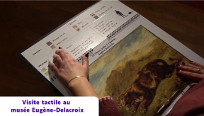 Gros plan sur une personne utilisant un livret de médiation tactile au musée Eugène-Delacroix. Ses mains parcourent des schémas en braille et une reproduction en relief du tableau « La Lionne prête à s'élancer ». Un bandeau indique : « Visite tactile au musée Eugène-Delacroix ».