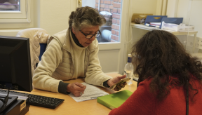 Au service social « La Bulle » (siège Duroc), une conseillère à lunettes montre un smartphone à une bénéficiaire de dos en pull rouge. Un échange administratif et numérique autour d'une table avec dossier et classeur vert.
