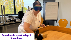 Une femme portant un masque occultant bleu et un gant de protection participe à une partie de Showdown lors de la Semaine du sport adapté. Elle est penchée au-dessus d'une table de jeu en bois rectangulaire avec des bords surélevés, tenant une raquette plate pour diriger une balle sonore. En arrière-plan, on aperçoit une salle lumineuse avec un tableau de conférence et des chaises en bois.