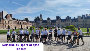 15 cyclistes portant des casques et des maillots de sport blancs posent pour une photo de groupe devant un immense château français historique, identifié comme le Château de Fontainebleau, sous un ciel bleu ensoleillé. Les participants se tiennent à côté de divers vélos, y compris des tandems visibles sur la droite, stationnés sur une allée pavée entourée de pelouses tondues et d'arbres taillés. Une bannière de texte dans un coin en bas à gauche contient l'inscription « Semaine du sport adapté Tandem »